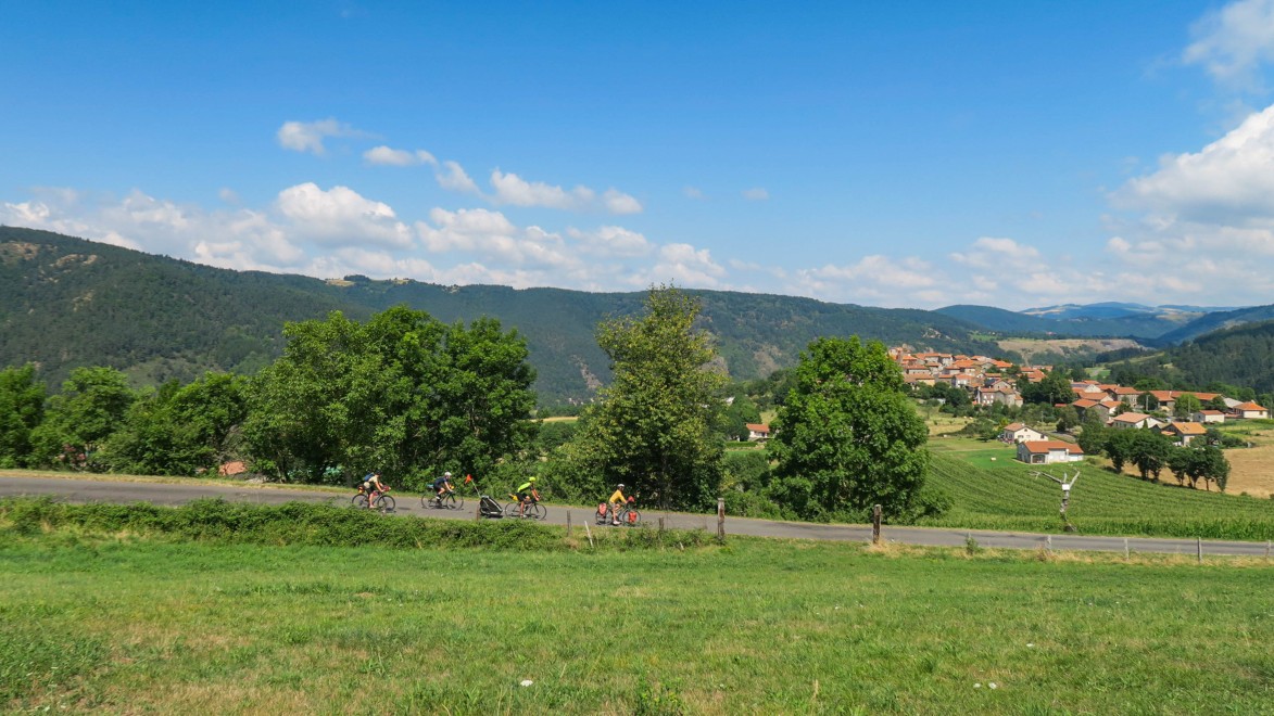 Vier Radfahrer in der Abfahrt in ein kleines Dorf im Allier, mit Wiesen und Wäldern bis zum Horizont.