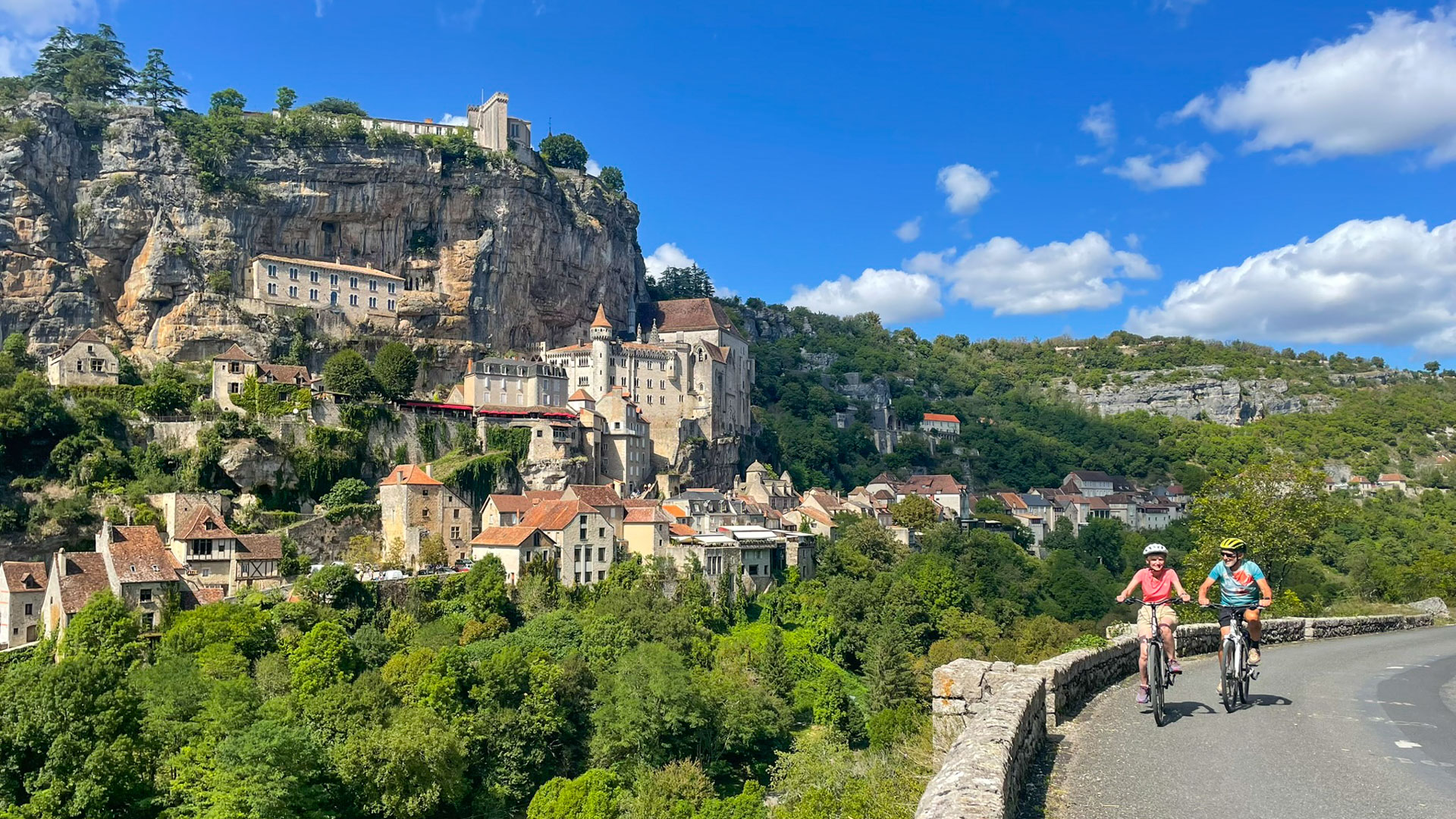 Ein Radfahrerpaar auf einer kleinen Straße mit der Wallfahrtskirche von Rocamadour im Hintergrund