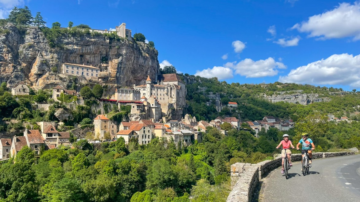 Ein Radfahrerpaar auf einer kleinen Straße mit der Wallfahrtskirche von Rocamadour im Hintergrund